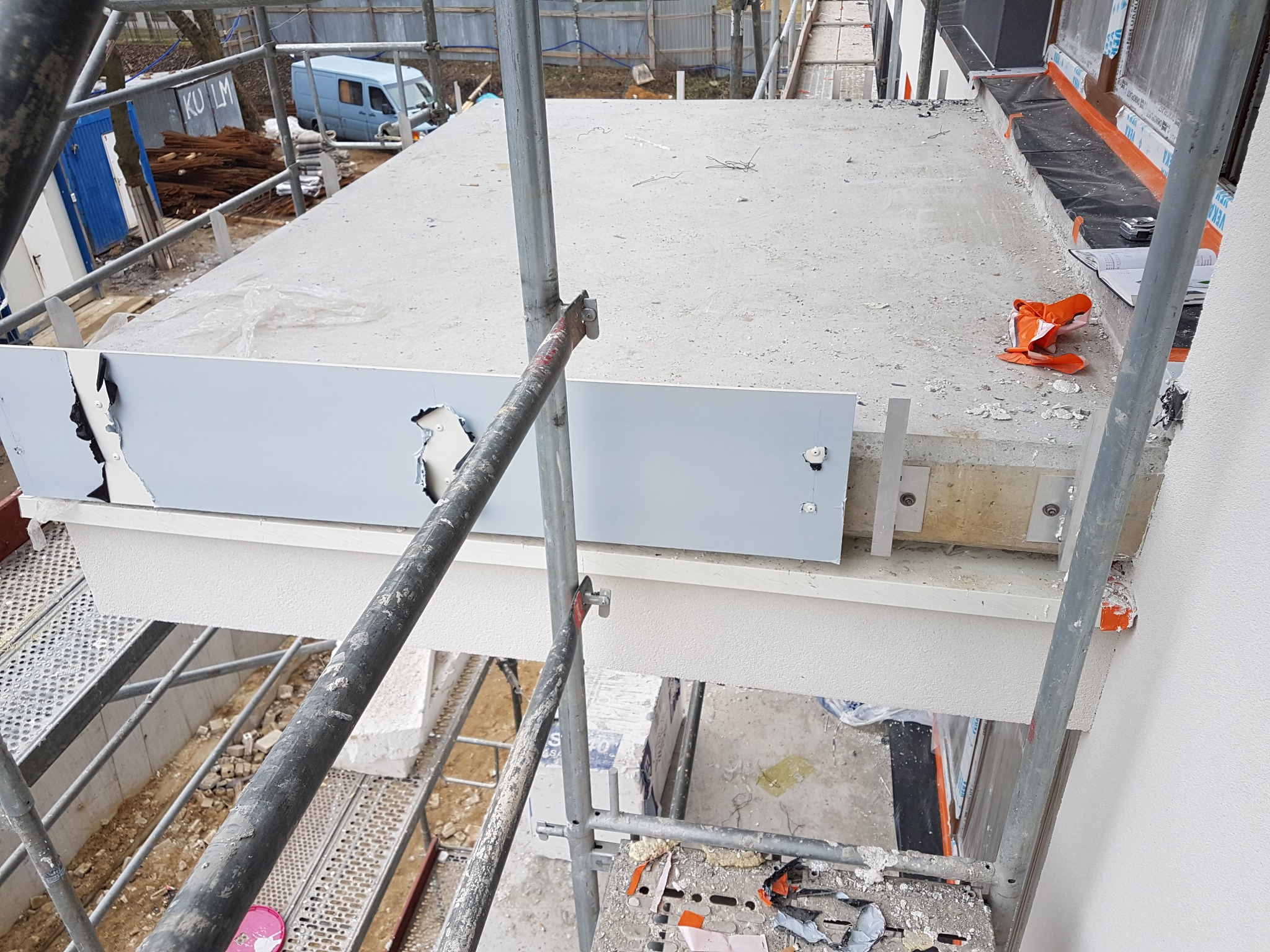 Widok z góry na fragment balkonu w trakcie budowy, widoczne rusztowanie, zabezpieczenie okna folią, elementy montażowe i uszkodzona płyta osłonowa.