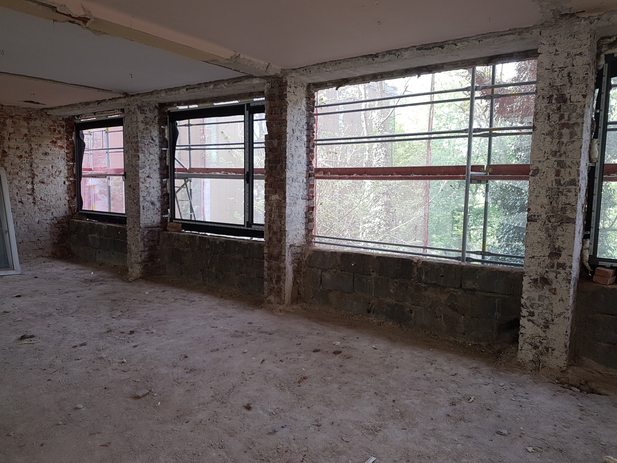 Wnętrze budynku w stanie surowym z trzema oknami; widoczne fragmenty cegieł i resztki tynku na ścianach; na zewnątrz rusztowania i zieleń.