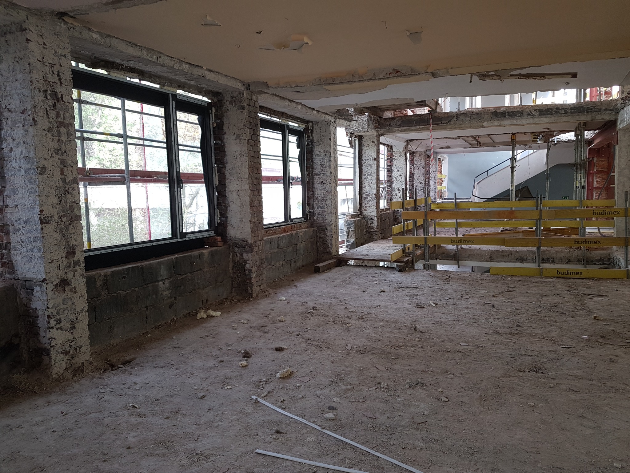 Wnętrze budynku w trakcie remontu, widoczne nowe okna z czarnymi ramami w surowych murach z cegły, podłoga pokryta gruzem, rusztowania w tle, częściowo odsłonięty sufit.
