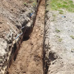 Długi, wąski wykop w ziemi, z widocznymi niebieskimi rurami i napiętym sznurkiem wyznaczającym linię, dwóch robotników w oddali, dom w tle.