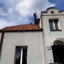 Mężczyzna na dachu budynku, w trakcie czyszczenia komina, trzyma w ręku szczotkę kominiarską, widok z dołu na tle błękitnego nieba z chmurami.