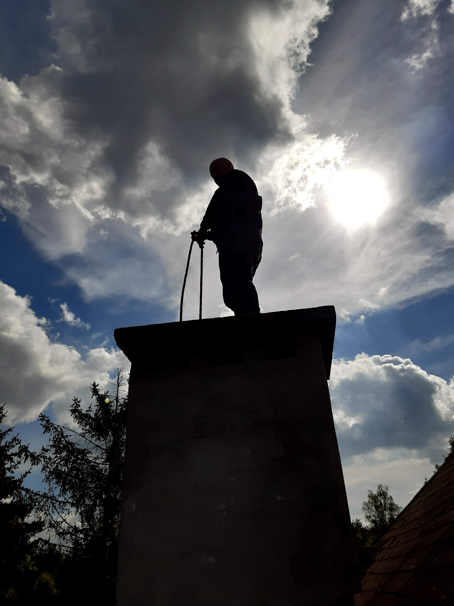 Sylwetka osoby stojącej na szczycie komina na tle pochmurnego nieba z prześwitującym słońcem, trzymającej w rękach narzędzia kominiarskie.