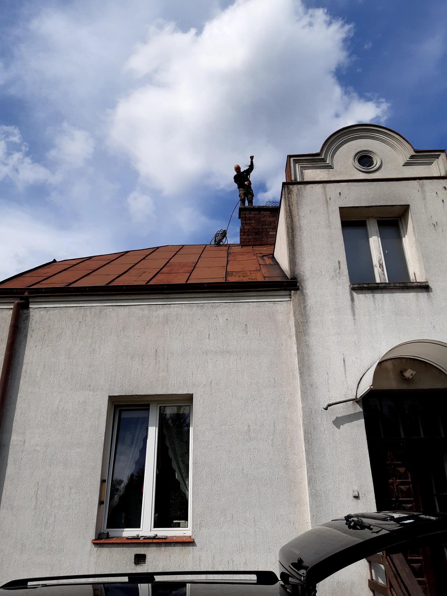 Mężczyzna na dachu budynku, w trakcie czyszczenia komina, trzyma w ręku szczotkę kominiarską, widok z dołu na tle błękitnego nieba z chmurami.