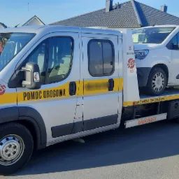 Srebrna laweta z napisem 'Pomoc Drogowa' przewozi białego busa na tle domów jednorodzinnych.