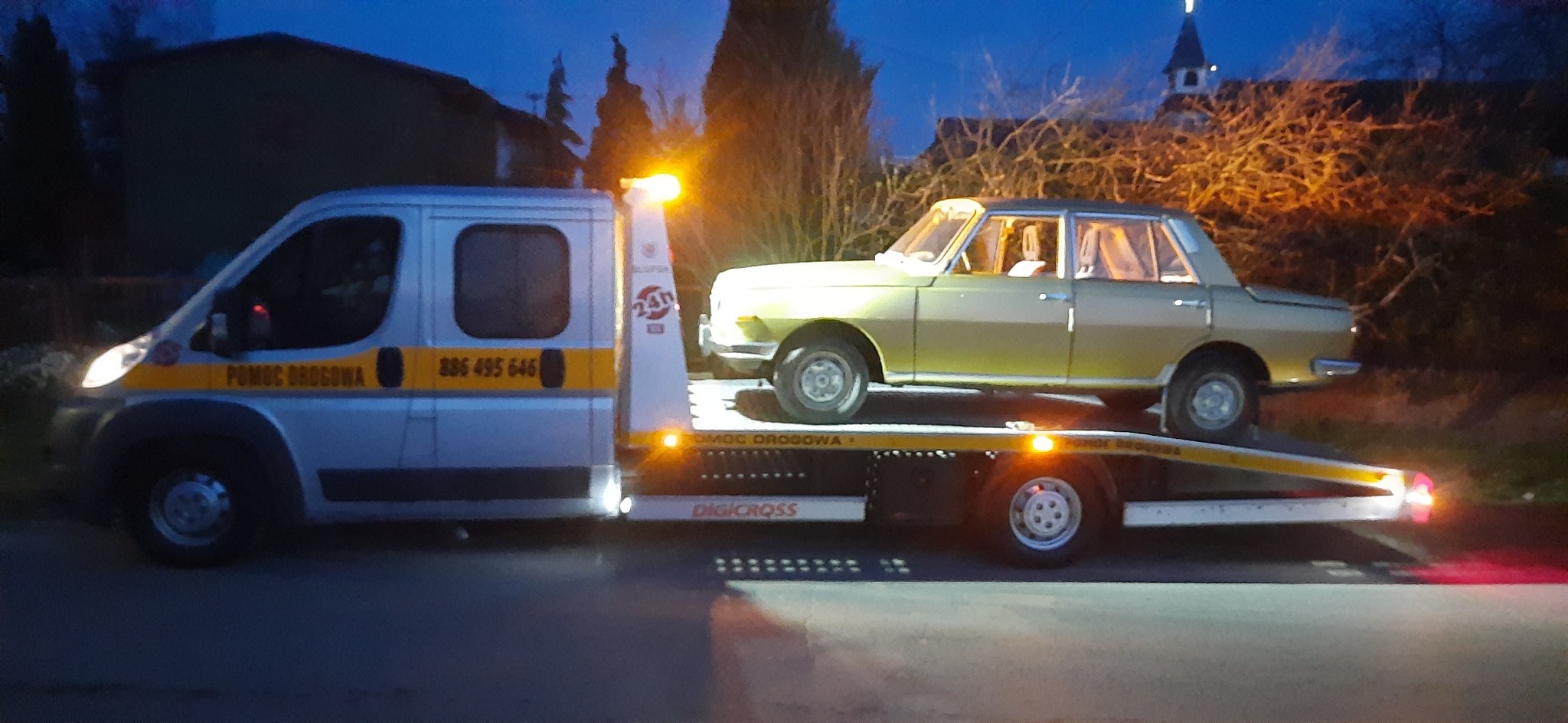 Srebrna laweta z włączonymi światłami ostrzegawczymi przewozi klasyczny, oliwkowy samochód osobowy na tle ciemnego, miejskiego krajobrazu z kościołem w oddali.