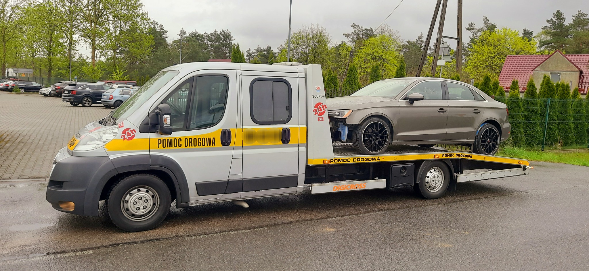 Szara laweta z uszkodzonym samochodem Audi A3 bez przedniego zderzaka, stojąca na parkingu w pochmurny dzień.