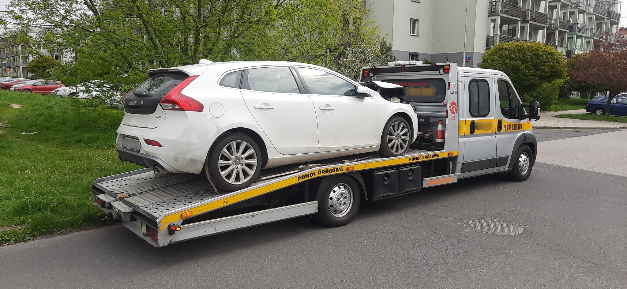 Biały uszkodzony samochód Volvo V40 na żółtej platformie lawety pomocy drogowej, na tle bloku mieszkalnego i zieleni.