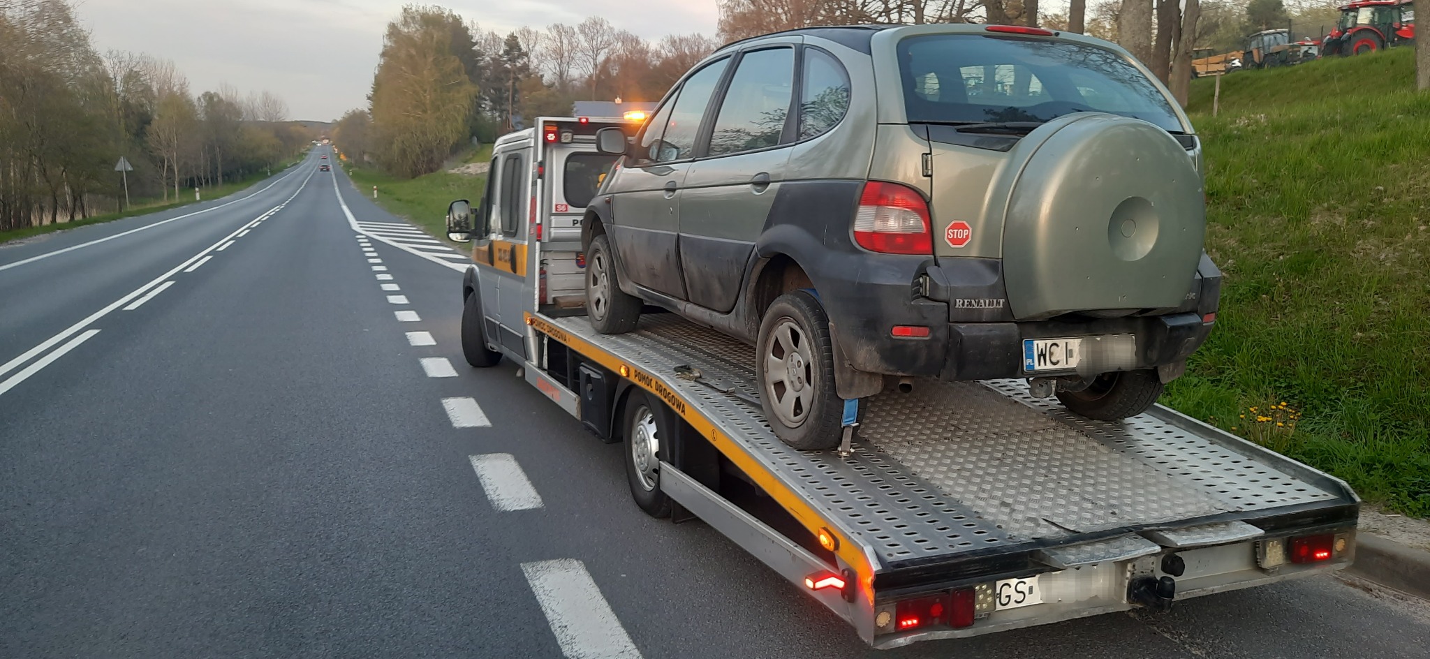 Samochód Renault Scenic zabezpieczony pasami na platformie pojazdu pomocy drogowej z włączonym pomarańczowym światłem ostrzegawczym, widok z tyłu w pochmurny dzień na drodze poza miastem.