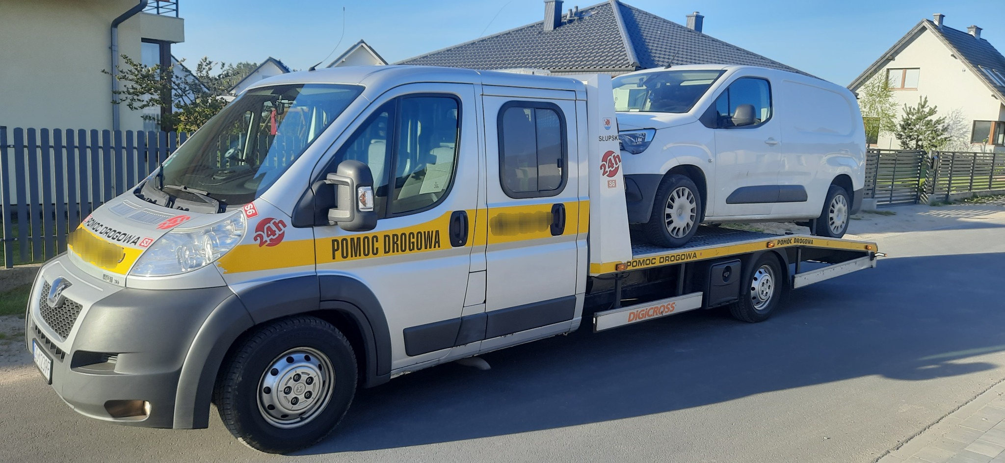 Srebrna laweta z napisem 'Pomoc Drogowa' przewozi białego busa na tle domów jednorodzinnych.