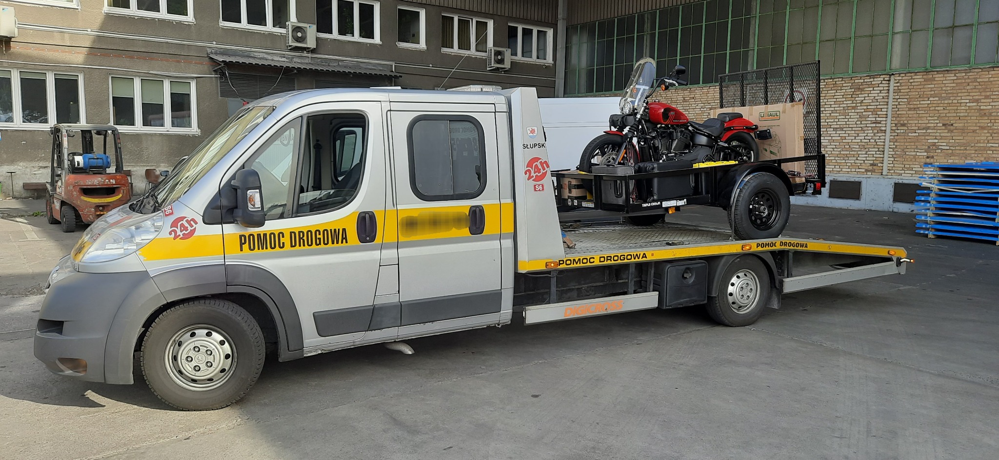 Srebrna laweta z żółtymi pasami 'POMOC DROGOWA' przewozi czerwony motocykl na przyczepie. W tle budynek przemysłowy i wózek widłowy.