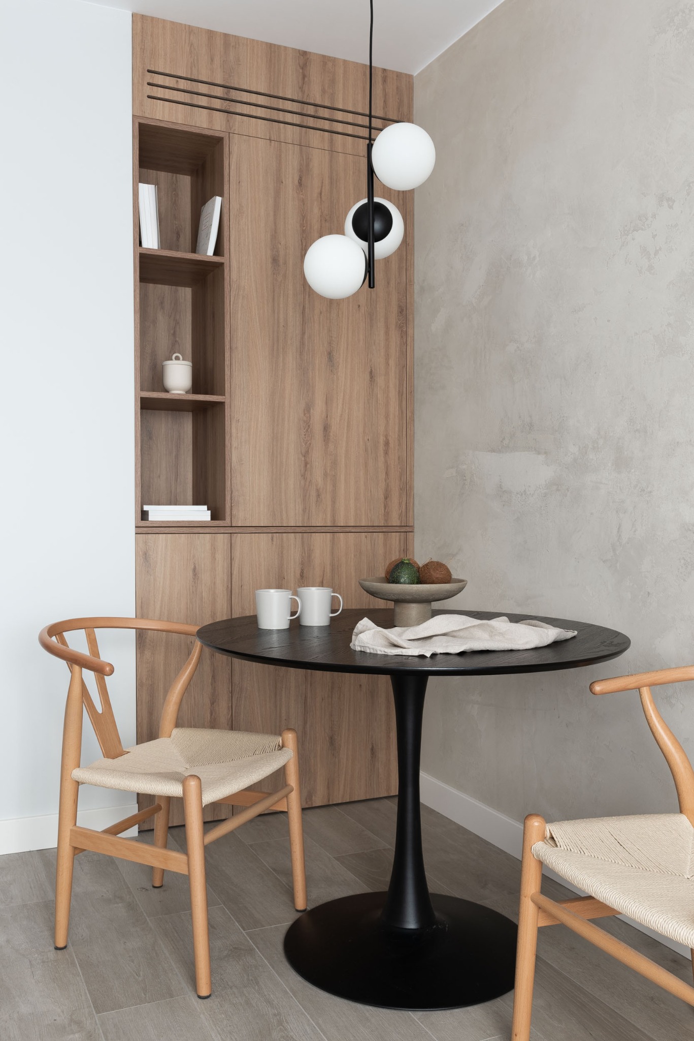 Nowoczesna aranżacja jadalni: okrągły, czarny stół na jednej nodze, dwie drewniane krzesła z jasnym siedziskiem, drewniana szafka z półkami, minimalistyczna lampa z kulistymi kloszami.
