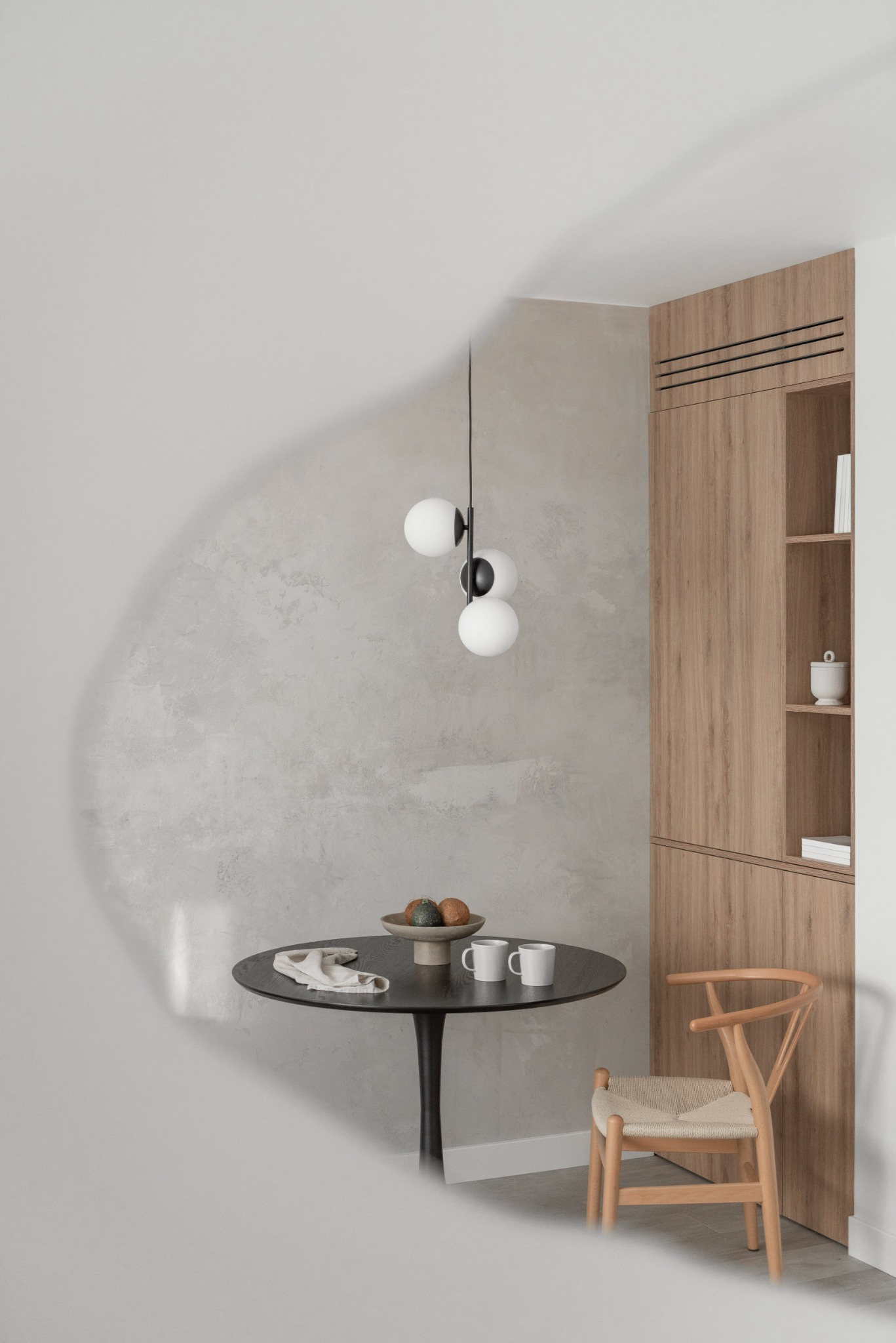 Minimalistyczna jadalnia z okrągłym czarnym stołem, dwiema białymi filiżankami, lnianą serwetką, owocami w misie, wiszącą lampą z trzema białymi kulami i drewnianą szafą w zabudowie oraz krzesłem...