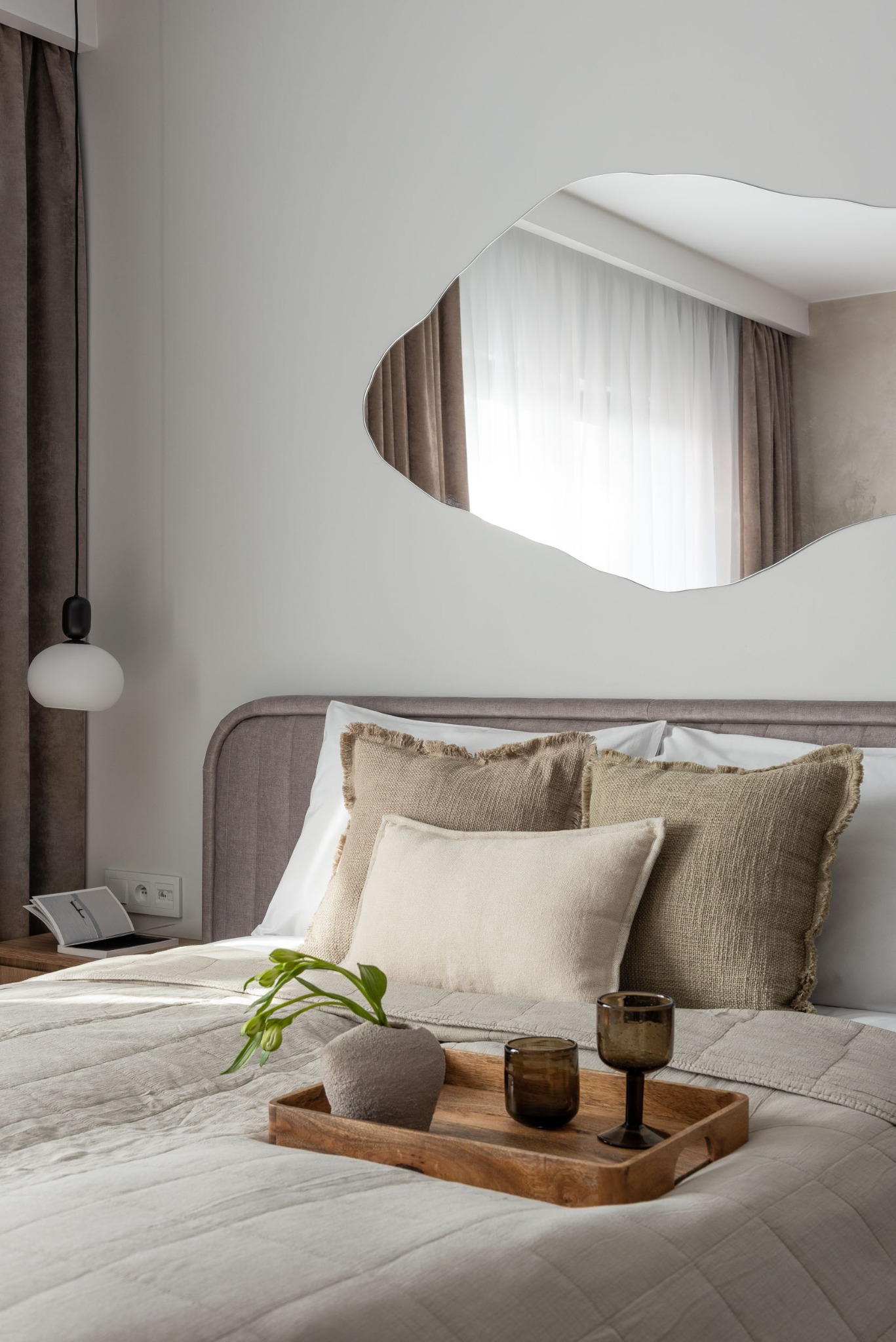 Neutralna sypialnia z łóżkiem, dekoracyjnymi poduszkami z frędzlami, asymetrycznym lustrem i drewnianą tacą z wazonem i kieliszkami.