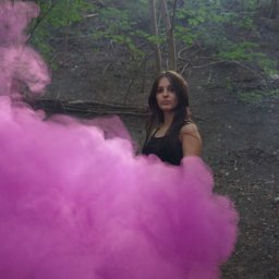 Klaudia Jadczak - Portret kobiety w lesie, otoczonej gęstym, różowym dymem, z ziemistym tłem i promieniami słońca przebijającymi przez drzewa.