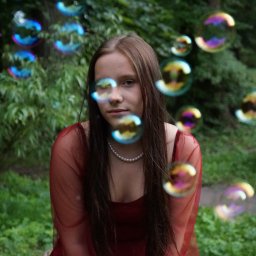 Klaudia Jadczak - Portret młodej kobiety w czerwonej sukience, otoczonej bańkami mydlanymi na tle zieleni. Delikatne światło, naturalna sceneria, perłowy naszyjnik.