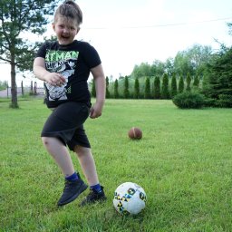 Klaudia Jadczak - Uśmiechnięty chłopiec w czarnej koszulce z motywem Batmana biegnie po trawie w kierunku piłki nożnej, w tle widoczne drzewa i krzewy.