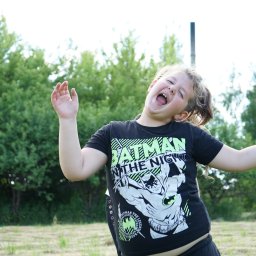 Klaudia Jadczak - Rozesmiana dziewczynka w czarnej koszulce z motywem Batmana, z szeroko otwartymi ustami i uniesionymi rękami, pozuje na tle zieleni.