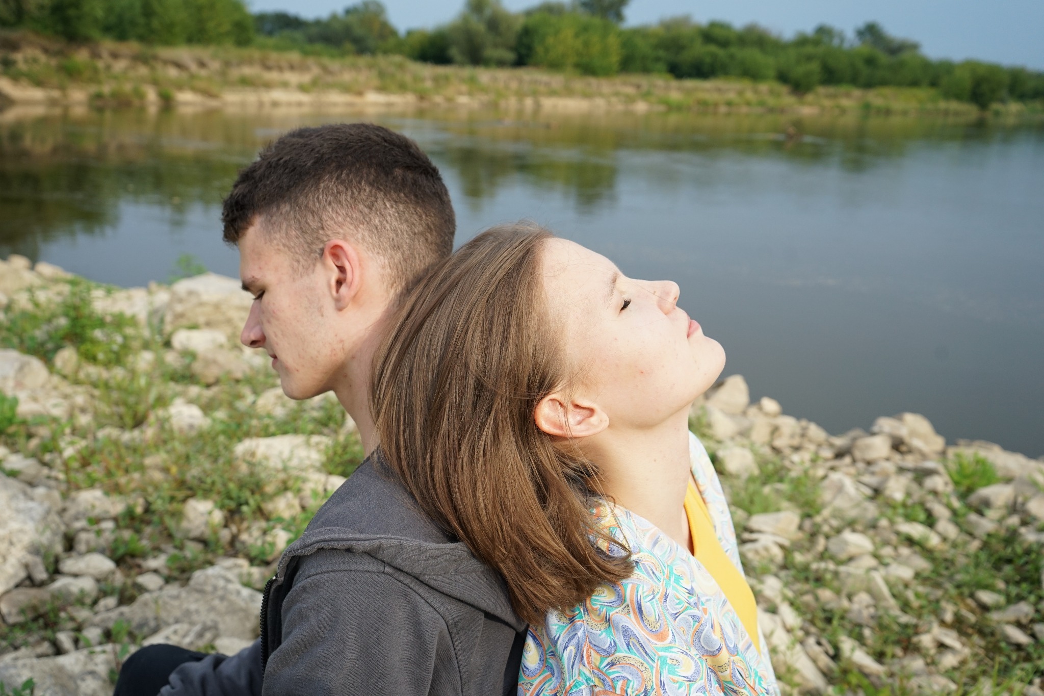 Para opiera się plecami na tle rzeki w słoneczny dzień: dziewczyna z głową zadartą do góry, chłopak ze spuszczonym wzrokiem.