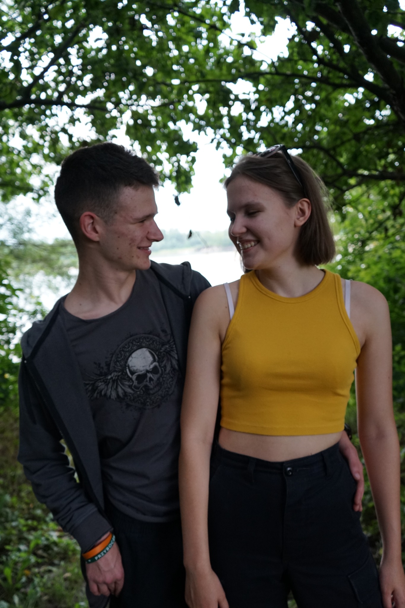 Uśmiechnięta para, mężczyzna w szarej bluzie z nadrukiem czaszki i kobieta w żółtym topie, stoją blisko siebie na tle zieleni drzew i jeziora w oddali.