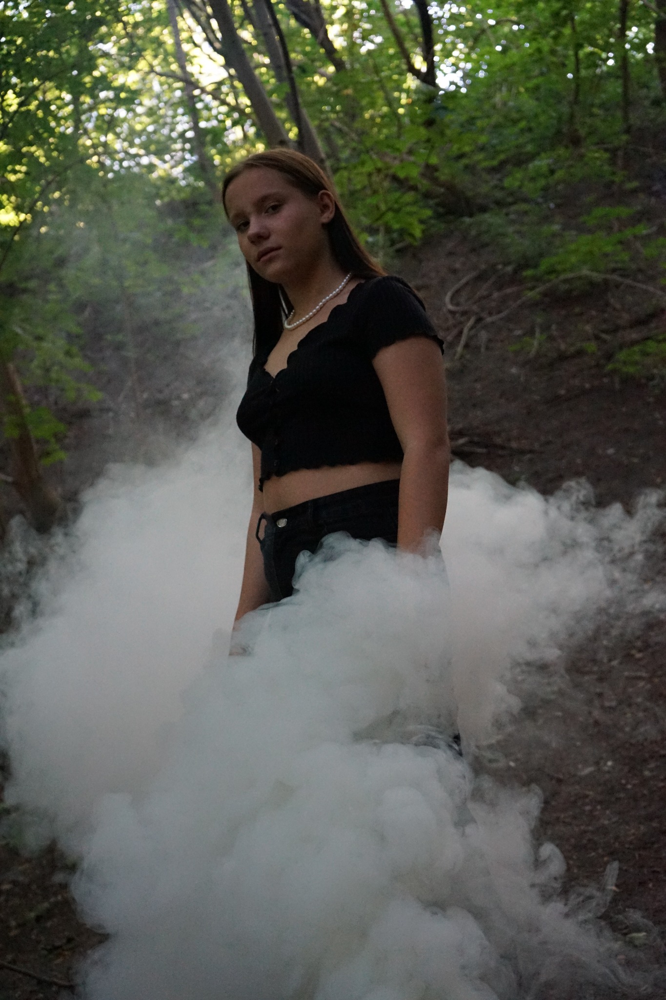 Portret kobiety w czarnym topie i perłach, otulonej dymem na tle leśnego zbocza w Warszawie.