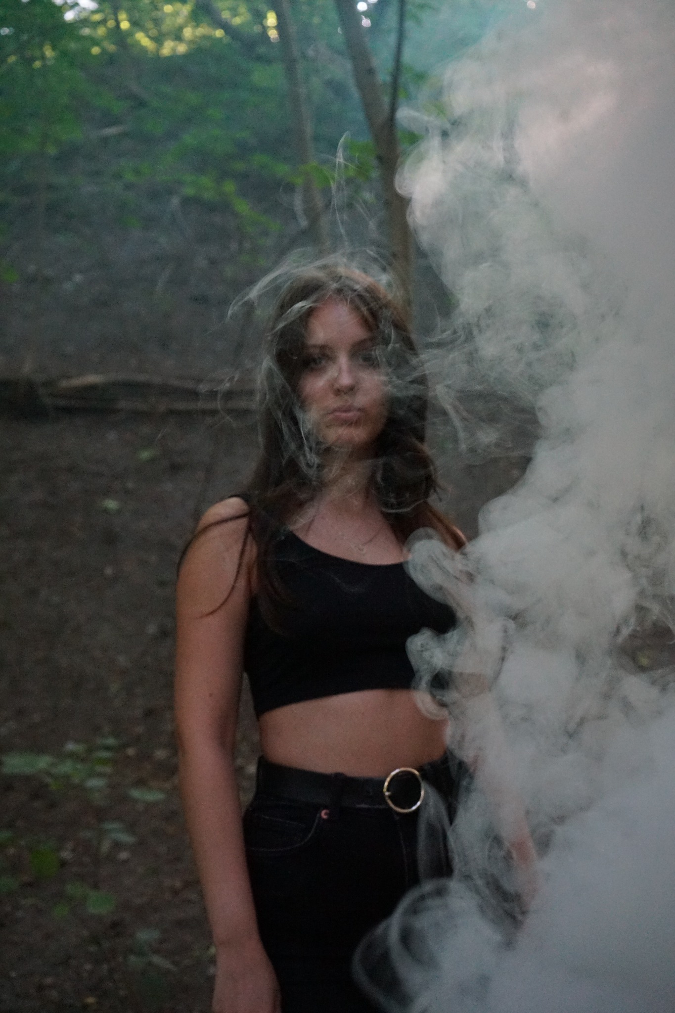 Portret kobiety w czarnym topie i dżinsach, otoczonej dymem w leśnym otoczeniu, z delikatnym makijażem i rozpuszczonymi włosami.