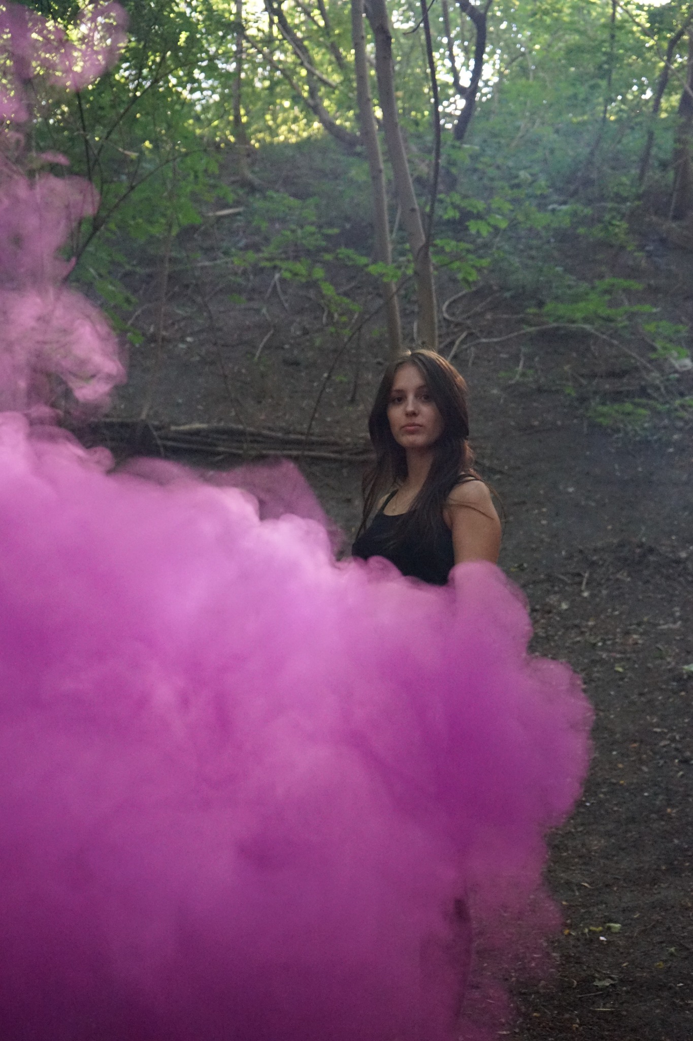Portret kobiety w lesie, otoczonej gęstym, różowym dymem, z ziemistym tłem i promieniami słońca przebijającymi przez drzewa.