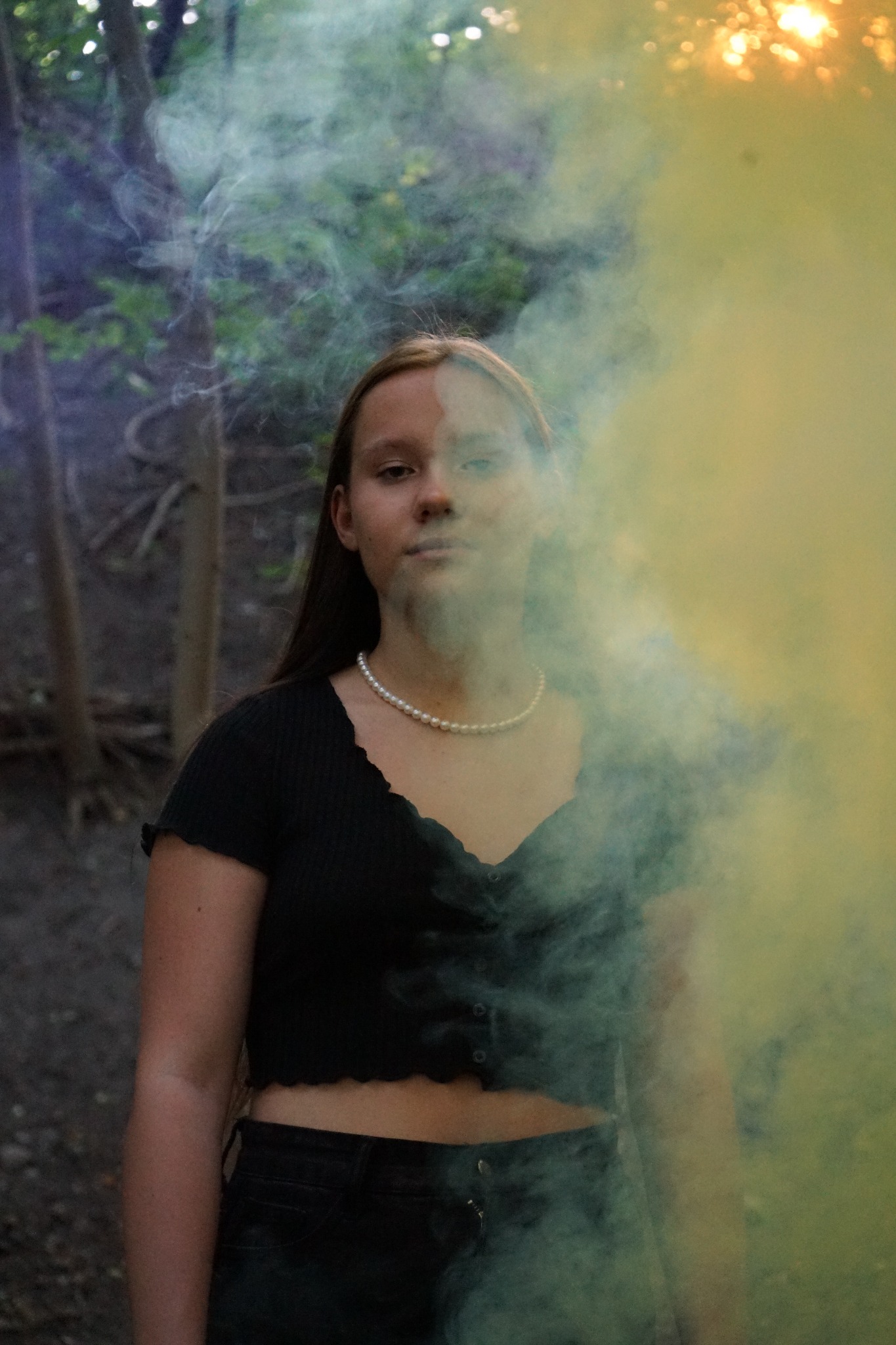 Młoda kobieta z perłowym naszyjnikiem pozuje w lesie, otoczona kolorowym dymem, z delikatnym oświetleniem słonecznym w tle.