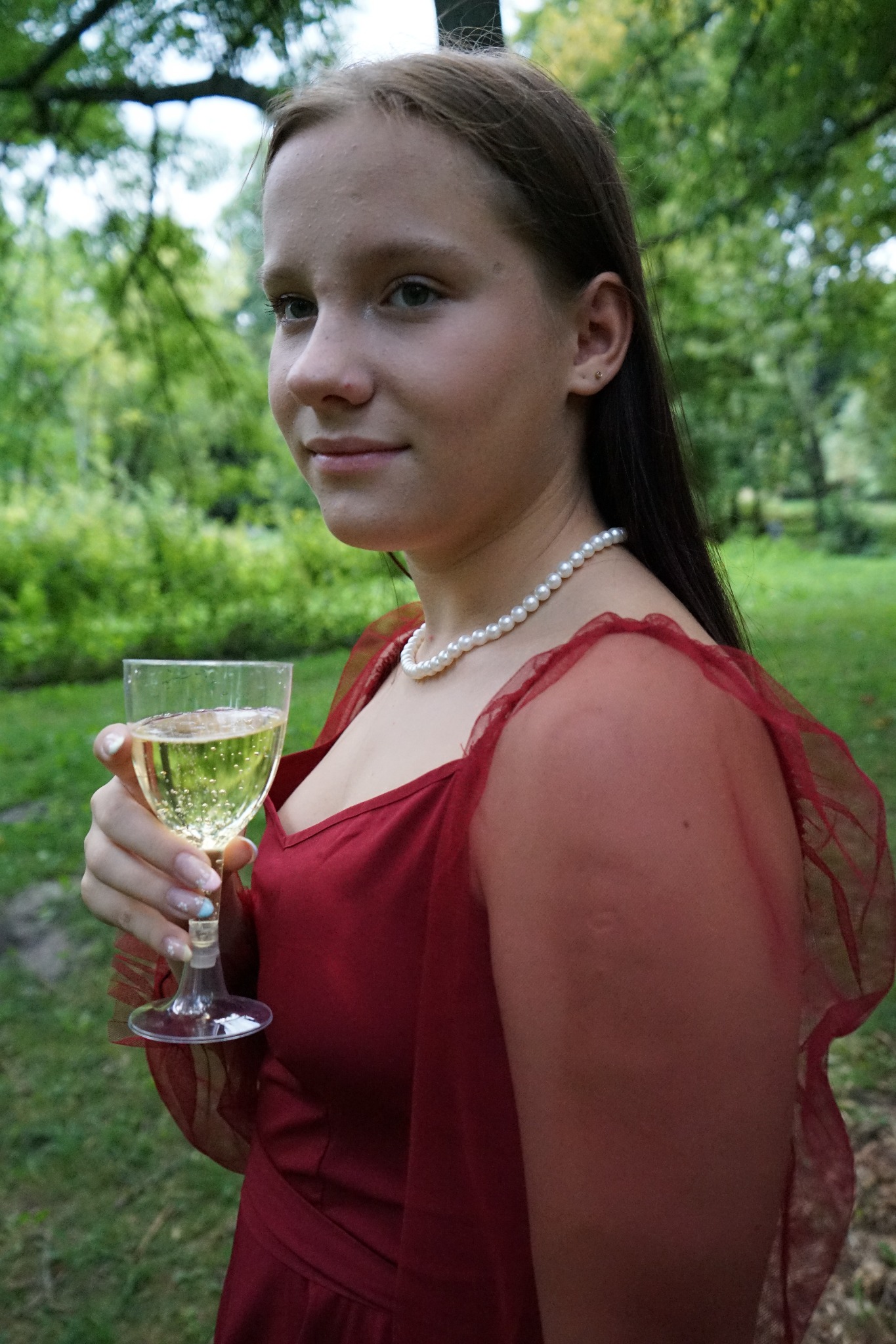 Młoda kobieta w czerwonej sukience i perłach, trzymająca kieliszek z musującym napojem na tle zielonego parku.