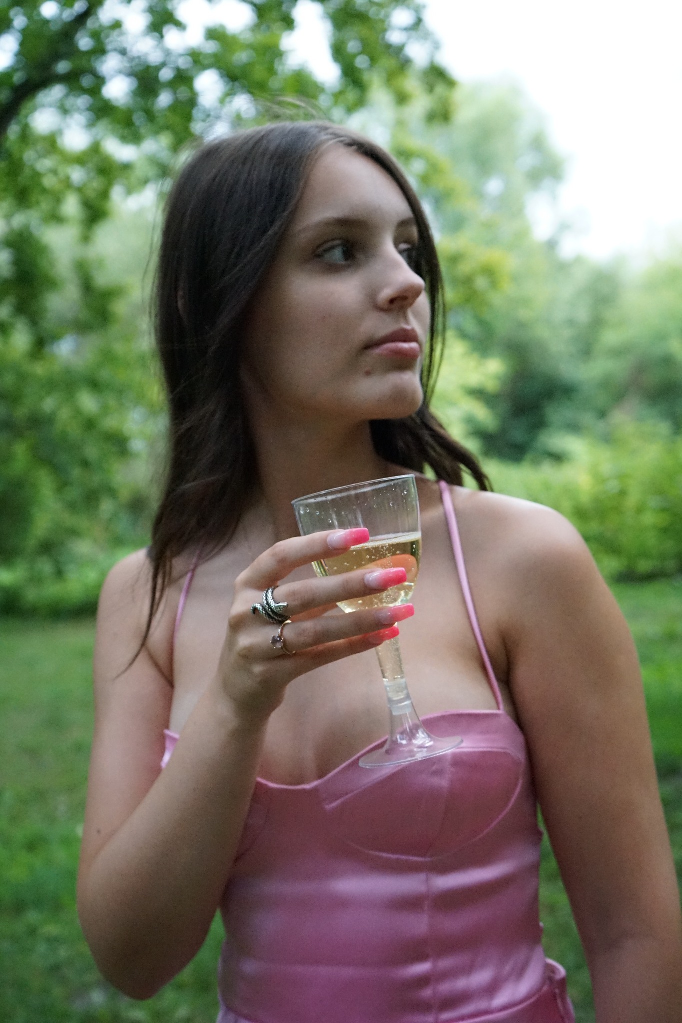 Młoda kobieta w różowej sukience, trzymająca kieliszek szampana na tle zieleni, z widocznymi pierścionkami na palcach i ombre manicure.