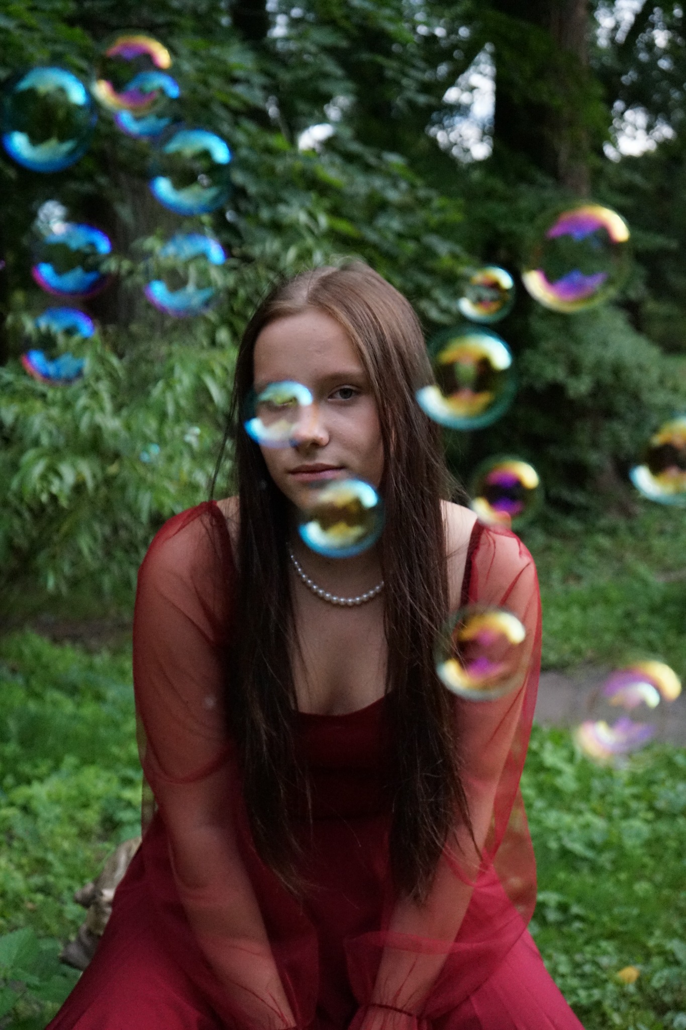 Portret młodej kobiety w czerwonej sukience, otoczonej bańkami mydlanymi na tle zieleni. Delikatne światło, naturalna sceneria, perłowy naszyjnik.