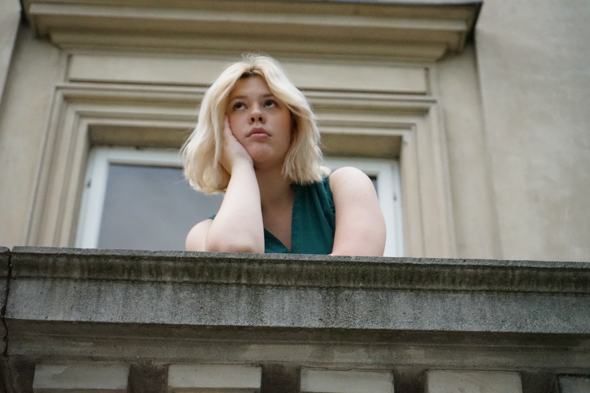 Portret młodej kobiety z blond włosami, opierającej się o kamienną balustradę balkonu, zamyślone spojrzenie skierowane w górę.