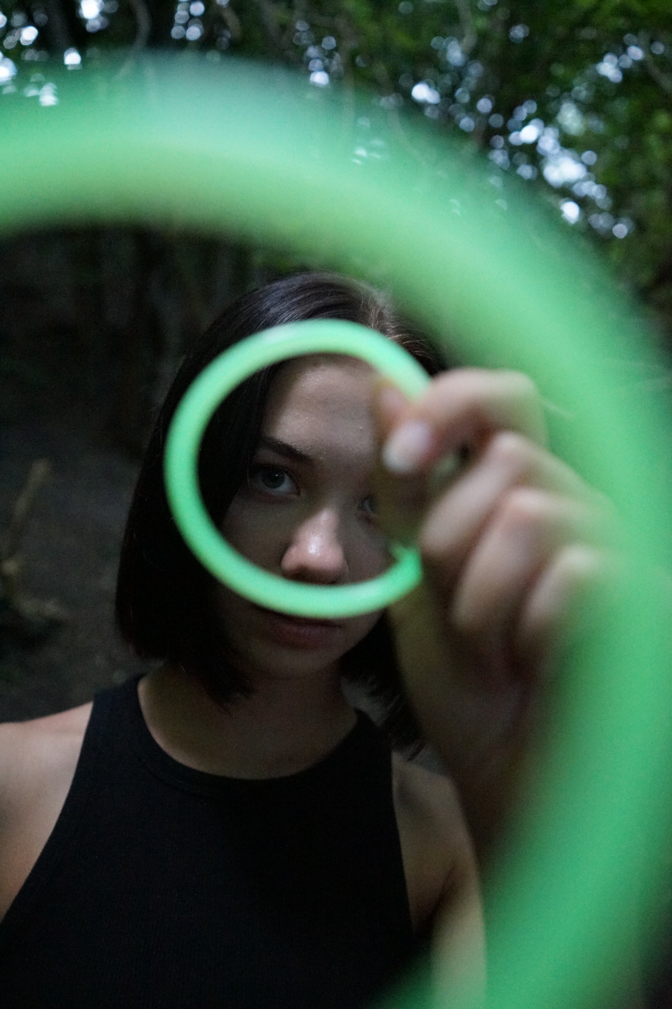 Portret młodej kobiety patrzącej przez neonowo-zielone kółko trzymane w dłoni, z ciemnym tłem leśnym w tle.