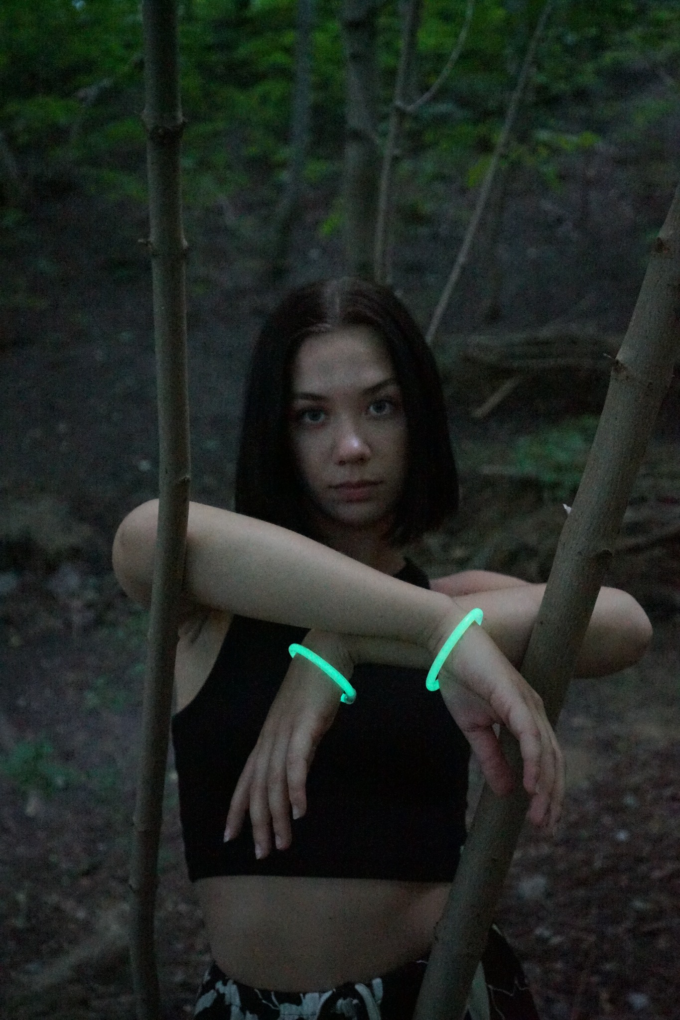 Portret młodej kobiety w lesie, z zielonymi, świecącymi bransoletkami na nadgarstkach, opierającej się o cienkie drzewka, w ciemnym oświetleniu.