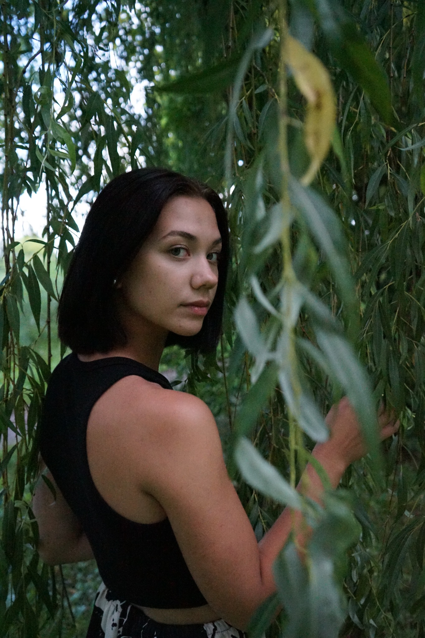Portret młodej kobiety z krótkimi, ciemnymi włosami, odwróconej przez ramię, na tle gęstych, zielonych gałęzi wierzby płaczącej.