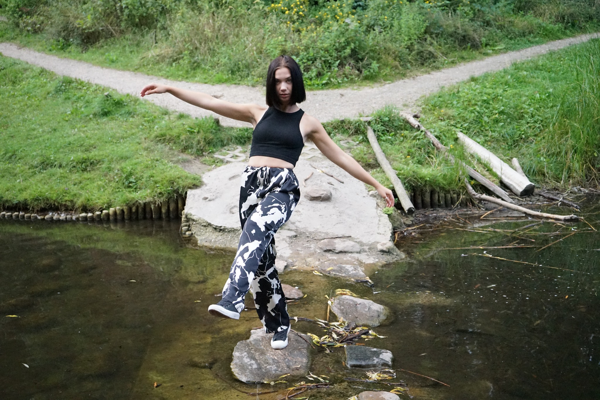 Młoda kobieta w czarnym topie i wzorzystych spodniach balansuje na kamieniach w płytkiej wodzie, z rozłożonymi ramionami, w tle rozwidlenie ścieżki i zielona roślinność.