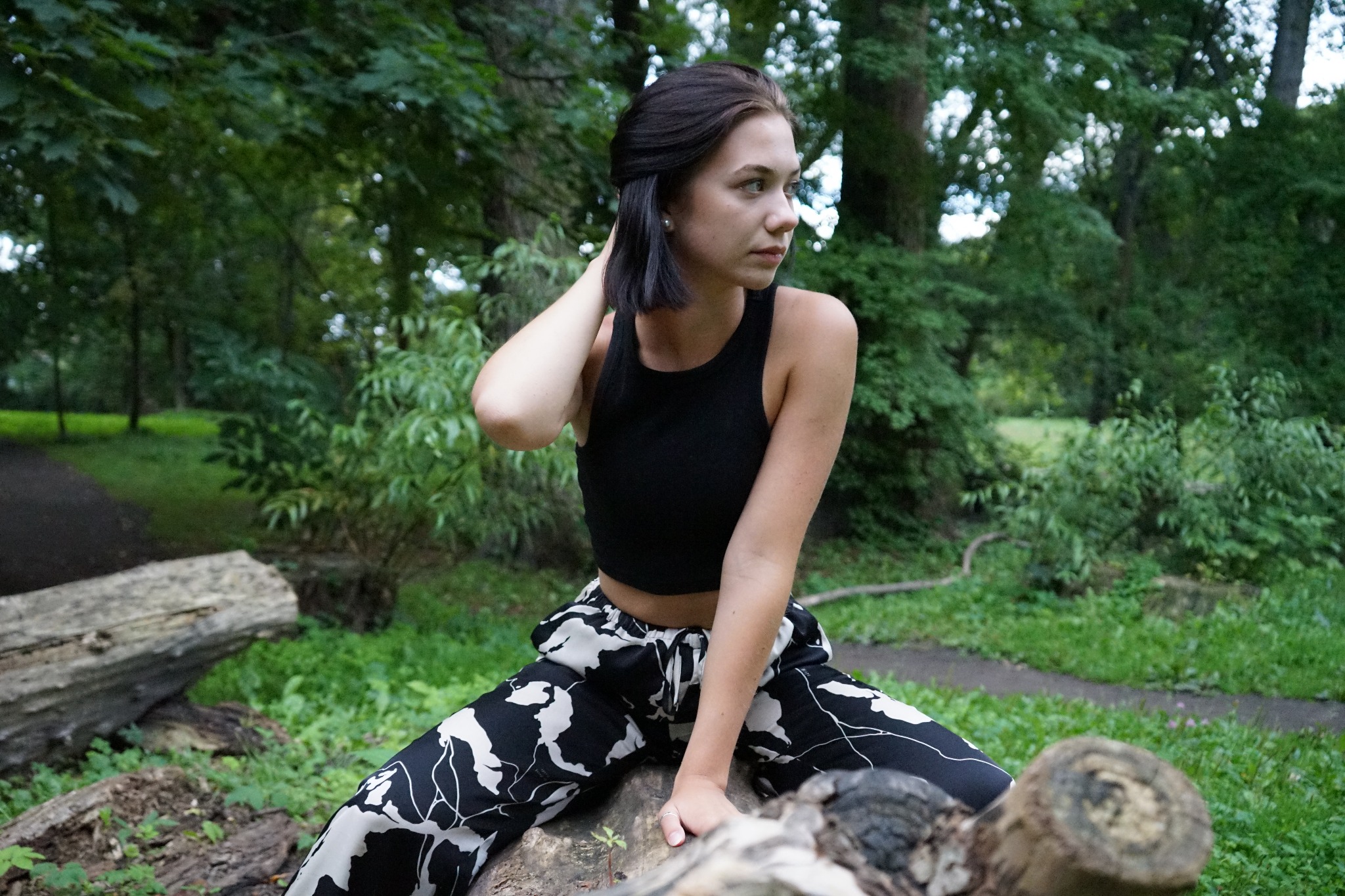 Kobieta w czarnym topie i wzorzystych spodniach siedzi na powalonym pniu drzewa w lesie, odwracając wzrok w prawo.