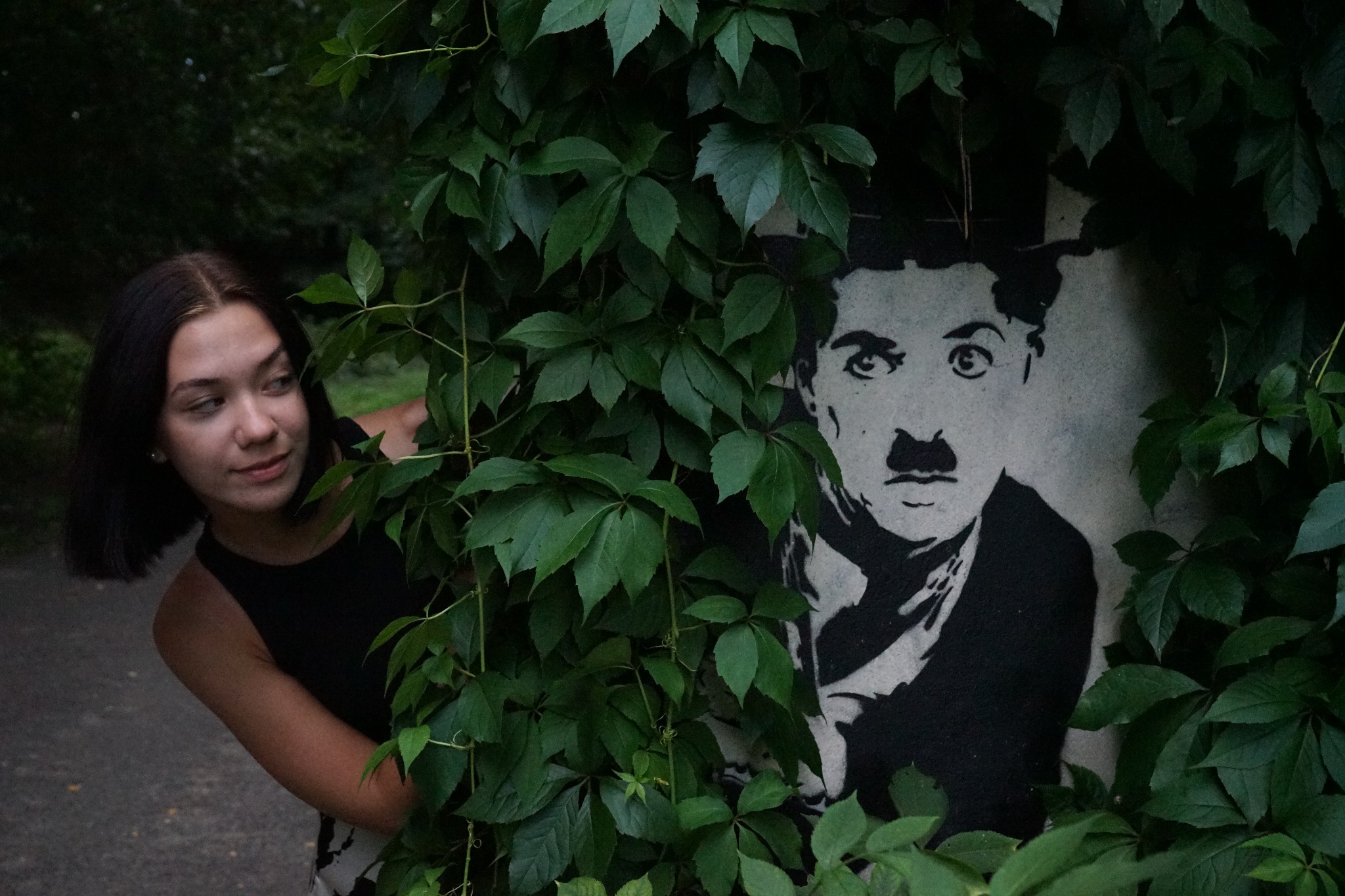 Młoda kobieta w czarnym topie wygląda zza gęstych zielonych liści, obok niej na ścianie widnieje czarno-biały portret Charlie Chaplina