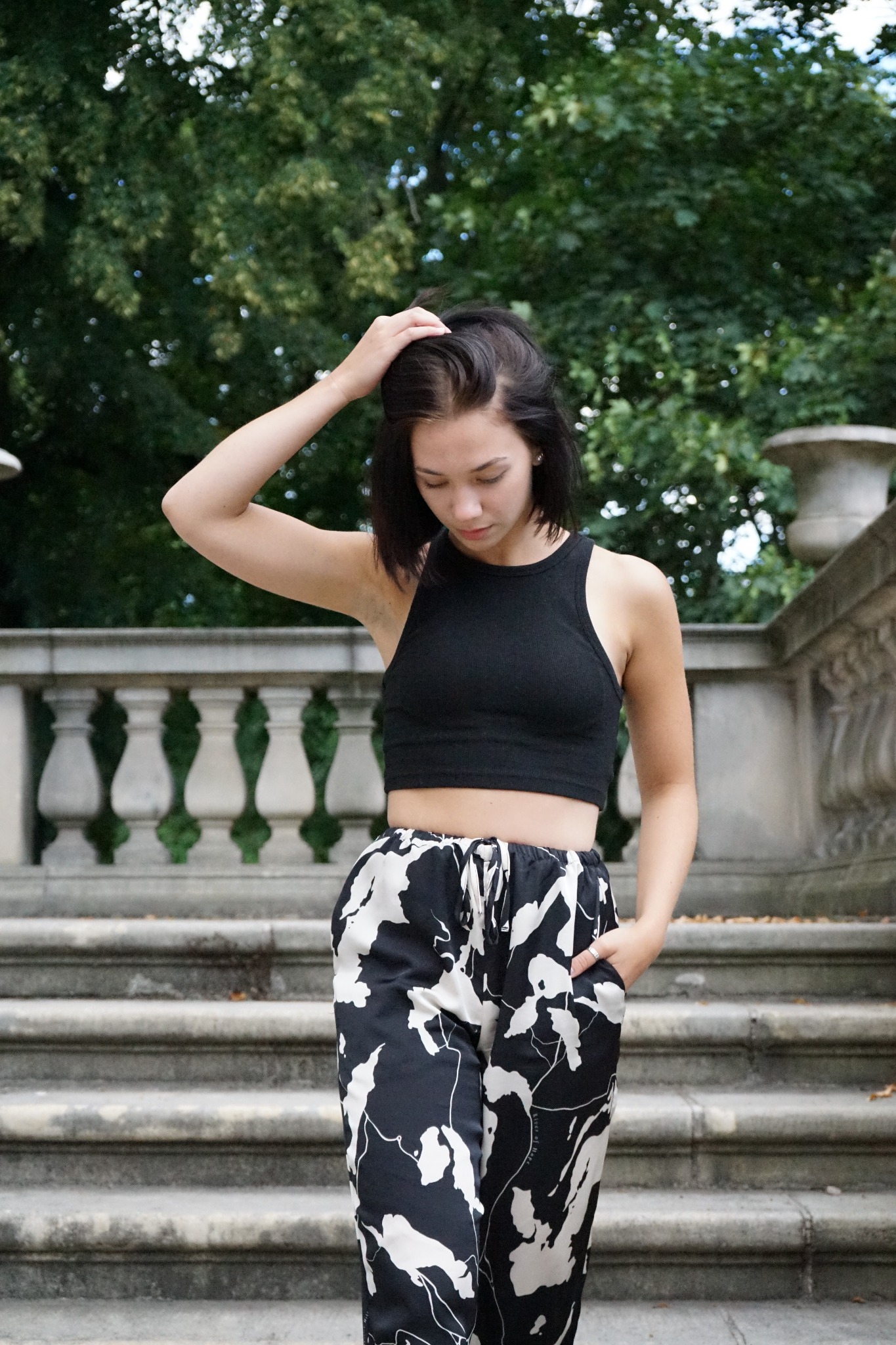Młoda kobieta w czarnym topie i wzorzystych spodniach, stojąca na kamiennych schodach z ręką we włosach i drugą w kieszeni, na tle zieleni.