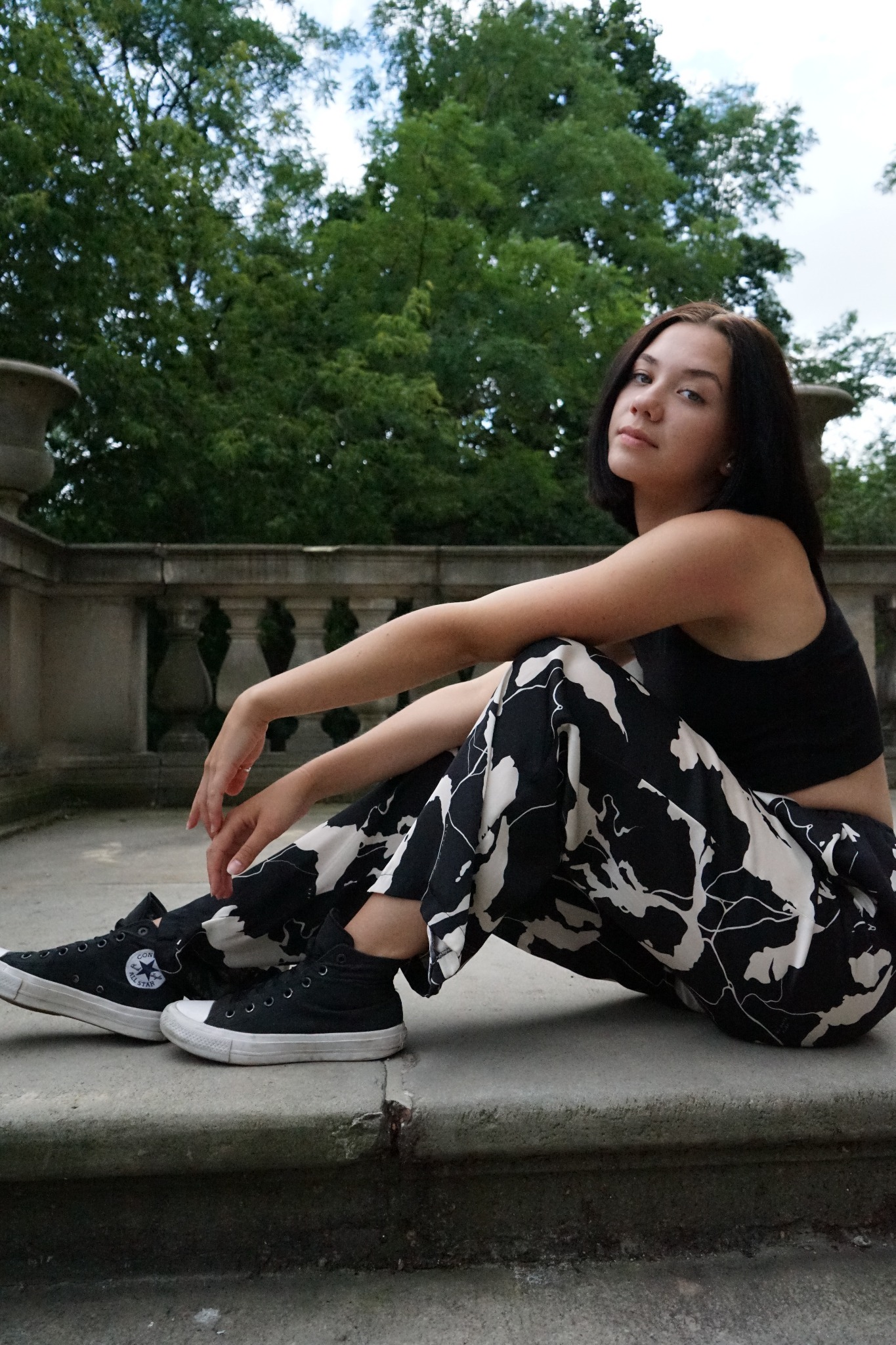 Portret młodej kobiety siedzącej na kamiennych schodach z balustradą w tle, ubrana w czarny top i wzorzyste spodnie, z butami Converse.