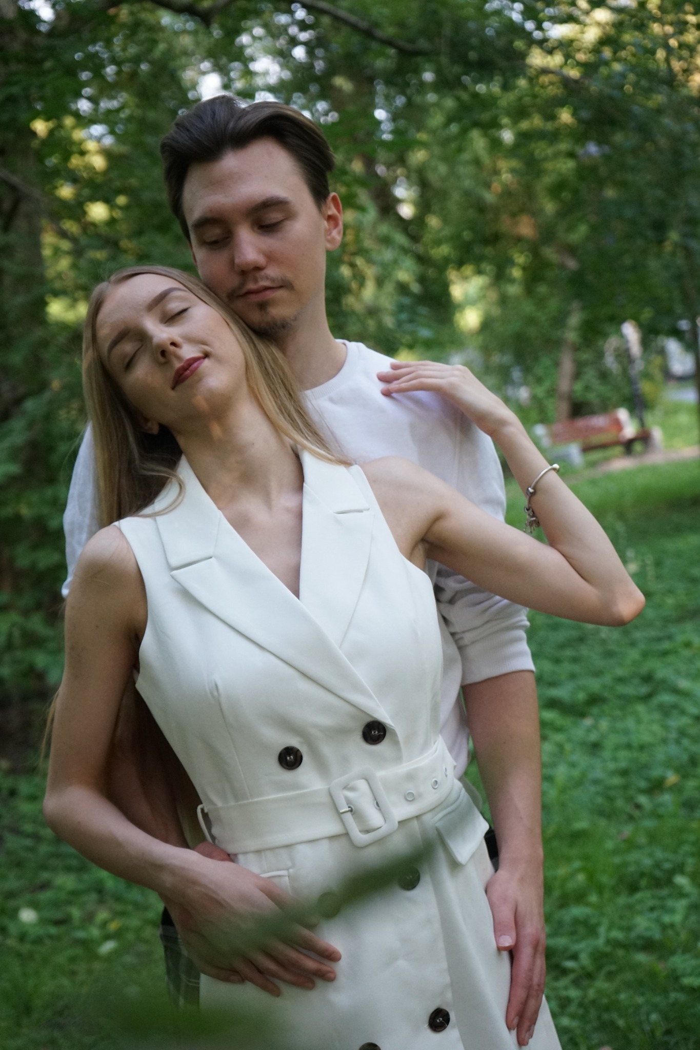 Młoda para w objęciach w parku; kobieta w białej sukience z paskiem, mężczyzna w białej koszulce, oboje z zamkniętymi oczami.