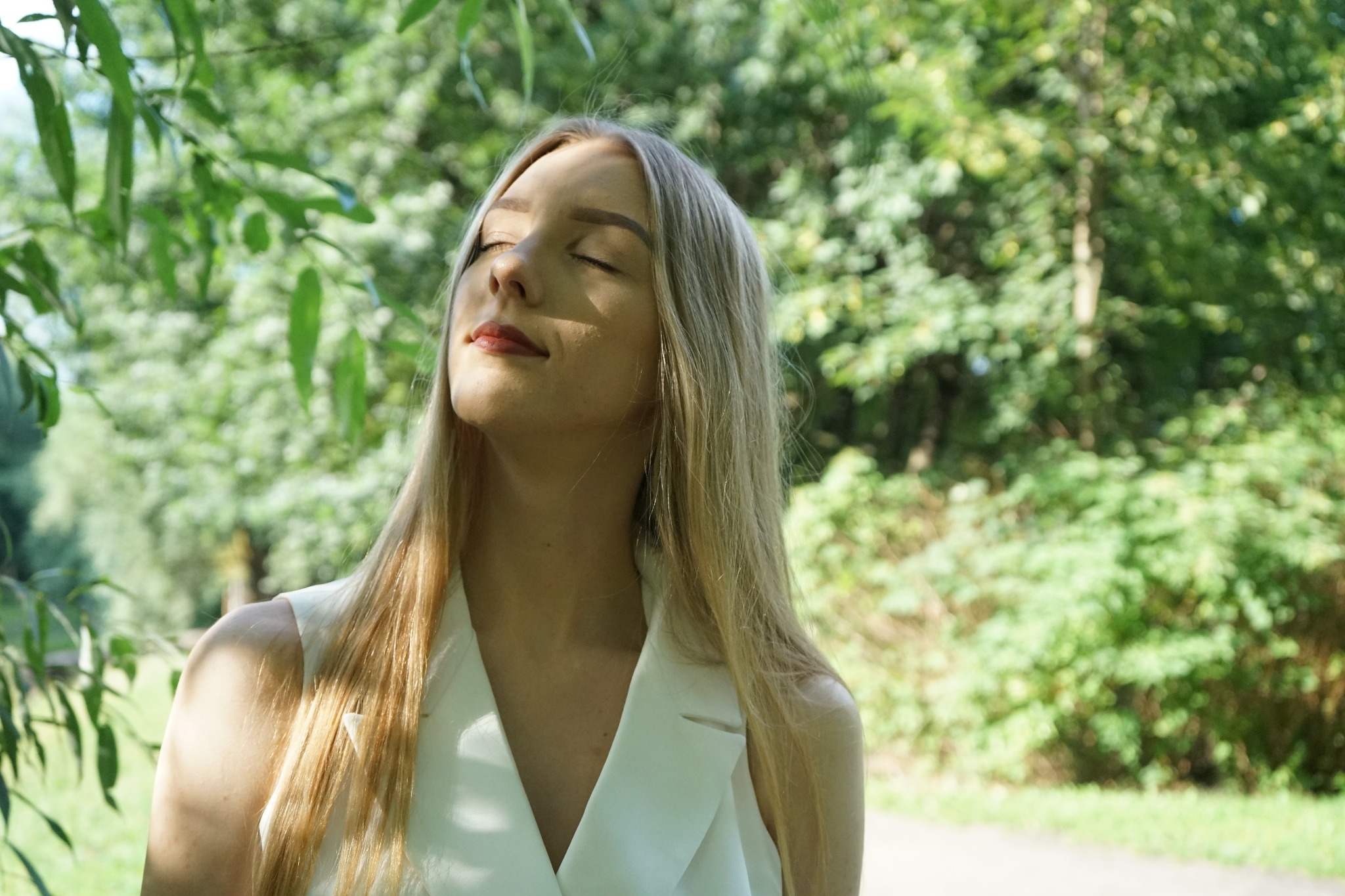 Portret kobiety z blond włosami i zamkniętymi oczami, w białej kamizelce na tle zieleni drzew, oświetlona naturalnym światłem.