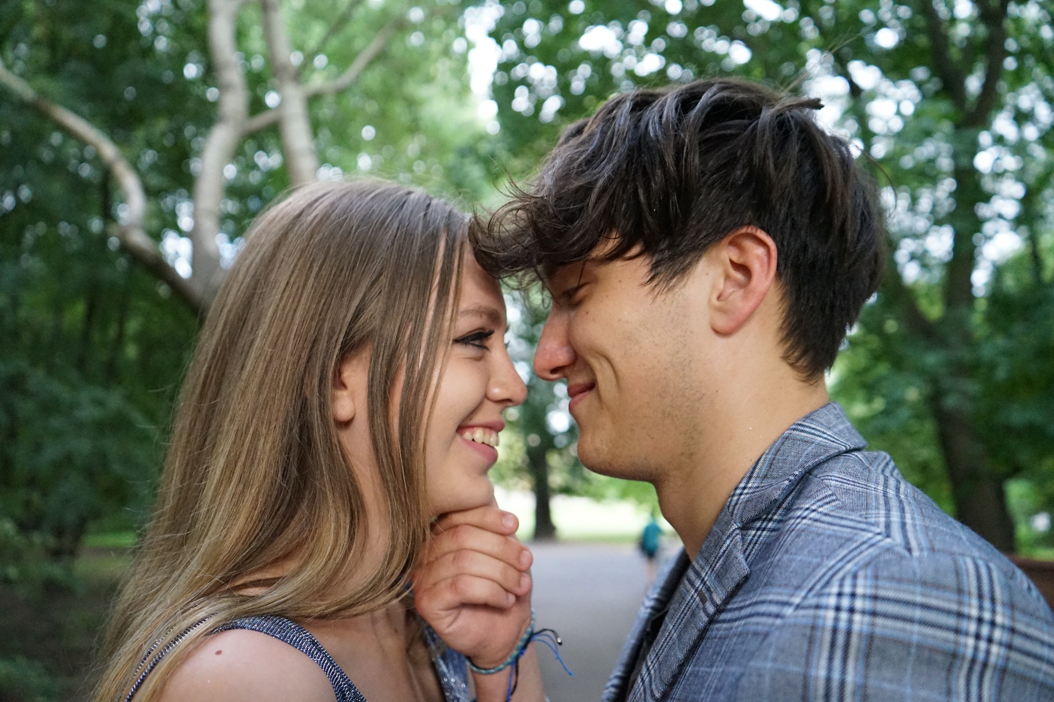 Uśmiechnięta para, stykając się czołami w plenerze, ona z blond włosami i niebieską bransoletką, on w szarej marynarce w kratę, drzewa w tle.