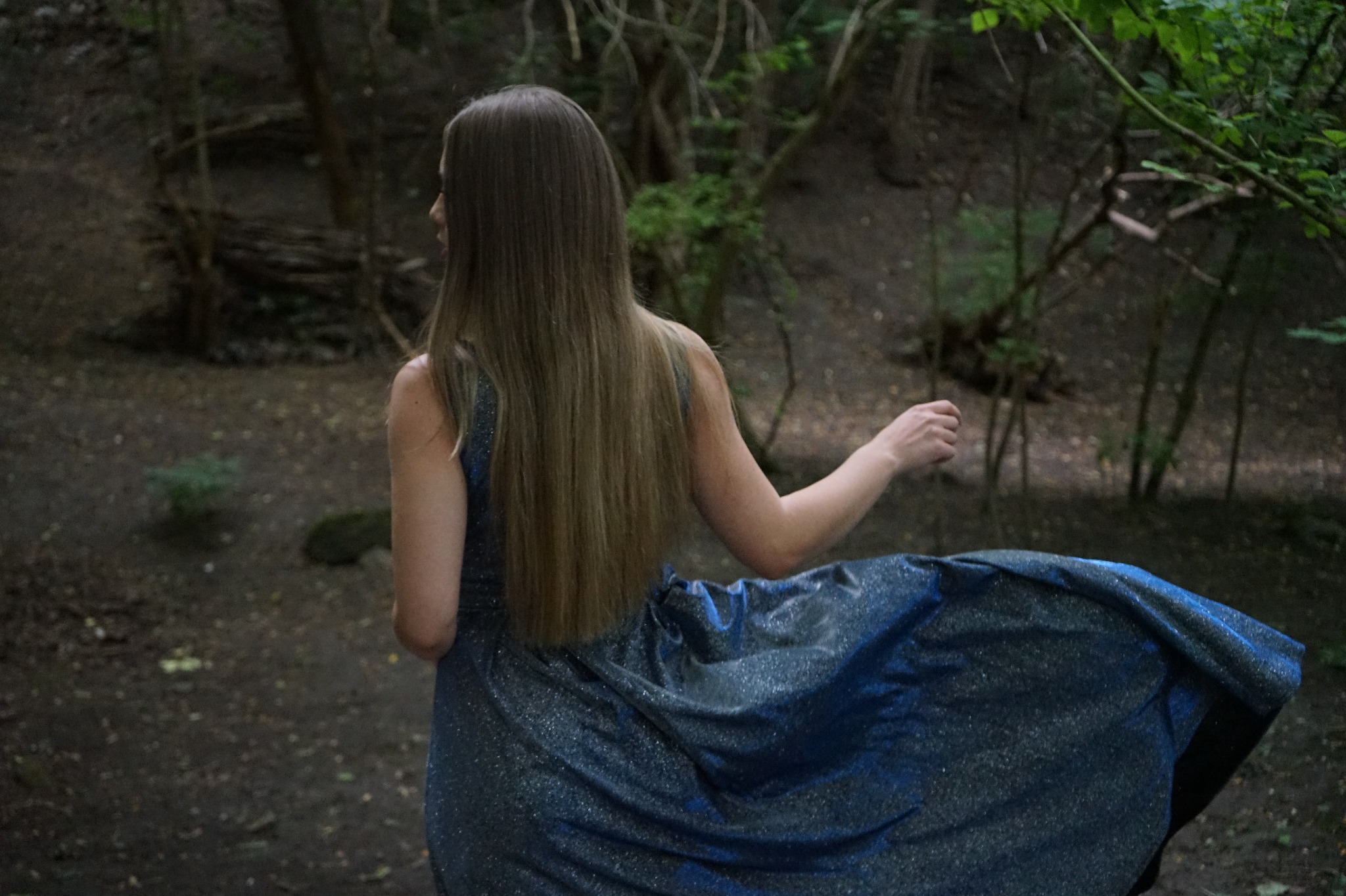 Kobieta w błyszczącej, niebieskiej sukni z odkrytymi plecami, sfotografowana od tyłu w leśnym otoczeniu, z rozłożoną spódnicą sukienki.