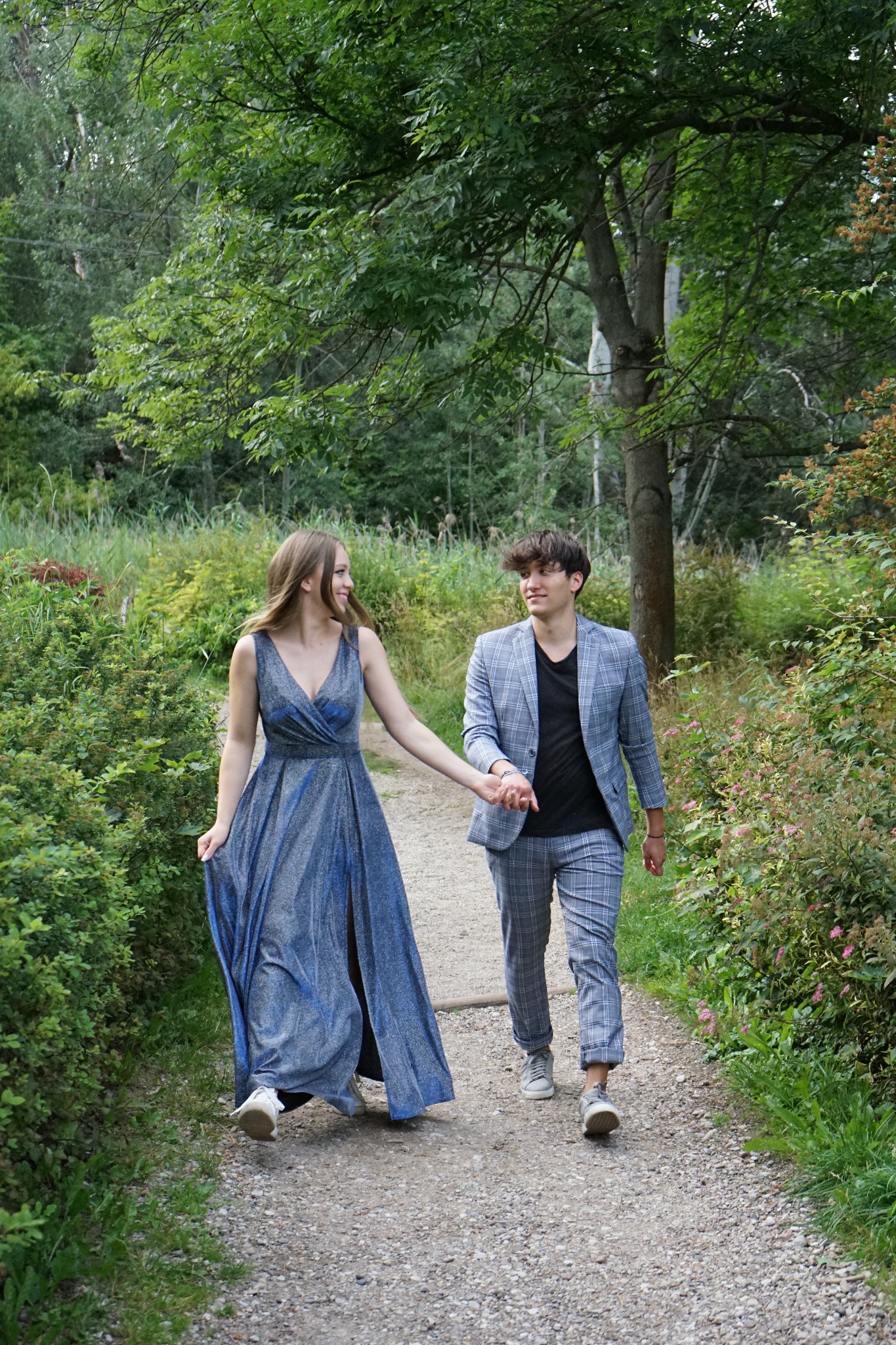 Para młodych spaceruje po leśnej ścieżce: ona w błyszczącej, niebieskiej sukni z rozcięciem, on w garniturze w kratę i czarnej koszulce.