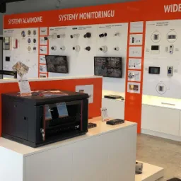 Ekspozycja systemów alarmowych i monitoringu w salonie: kamery, domofony, szafa rack z osprzętem na białym postumencie.