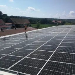 Montaż paneli fotowoltaicznych na dachu budynku gospodarczego z widocznymi pracownikami w oddali, otoczonym zielenią pól i lasów pod błękitnym niebem.