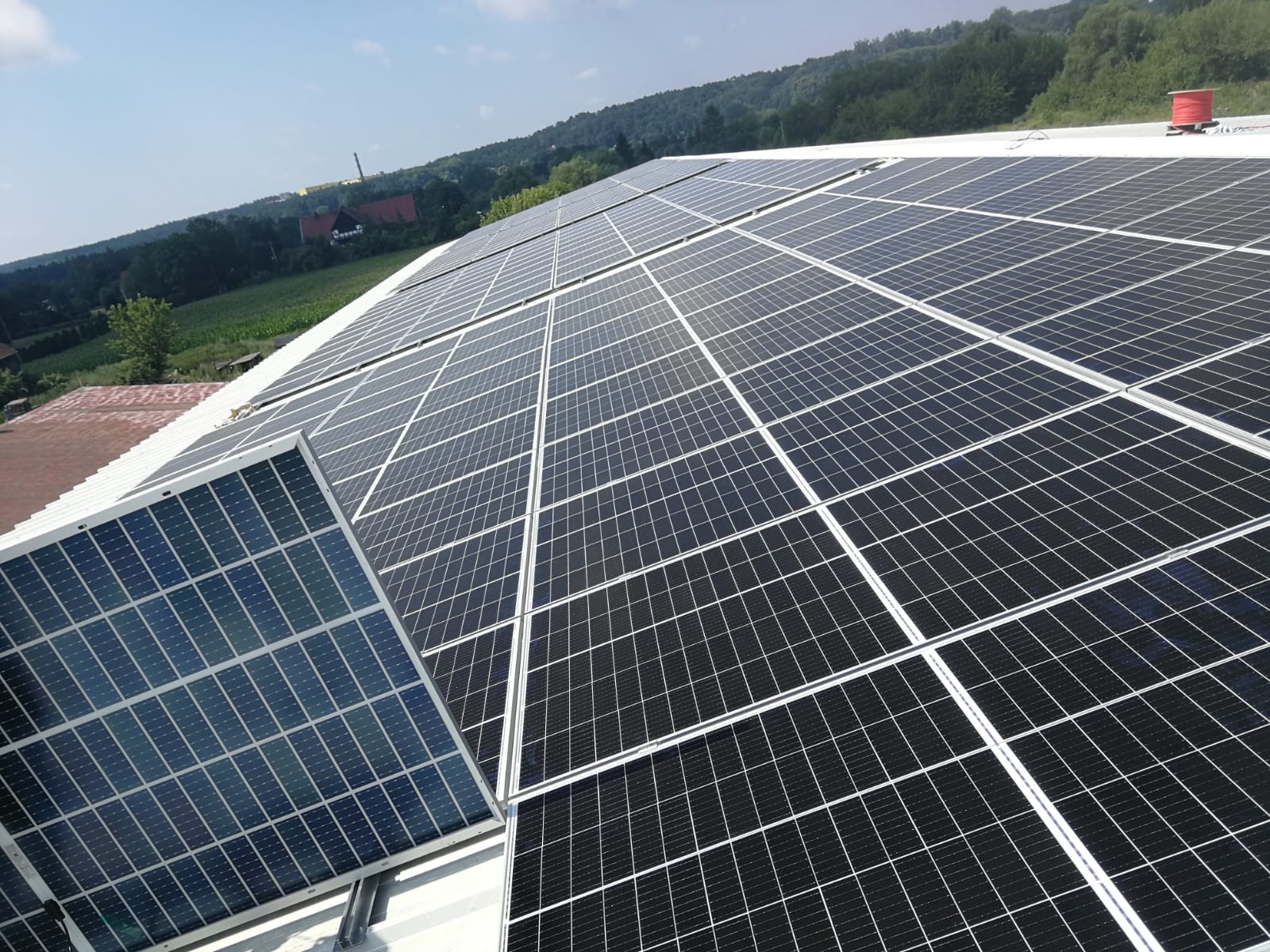 Rozległa instalacja fotowoltaiczna na dachu budynku z widokiem na zalesione wzgórza w tle.