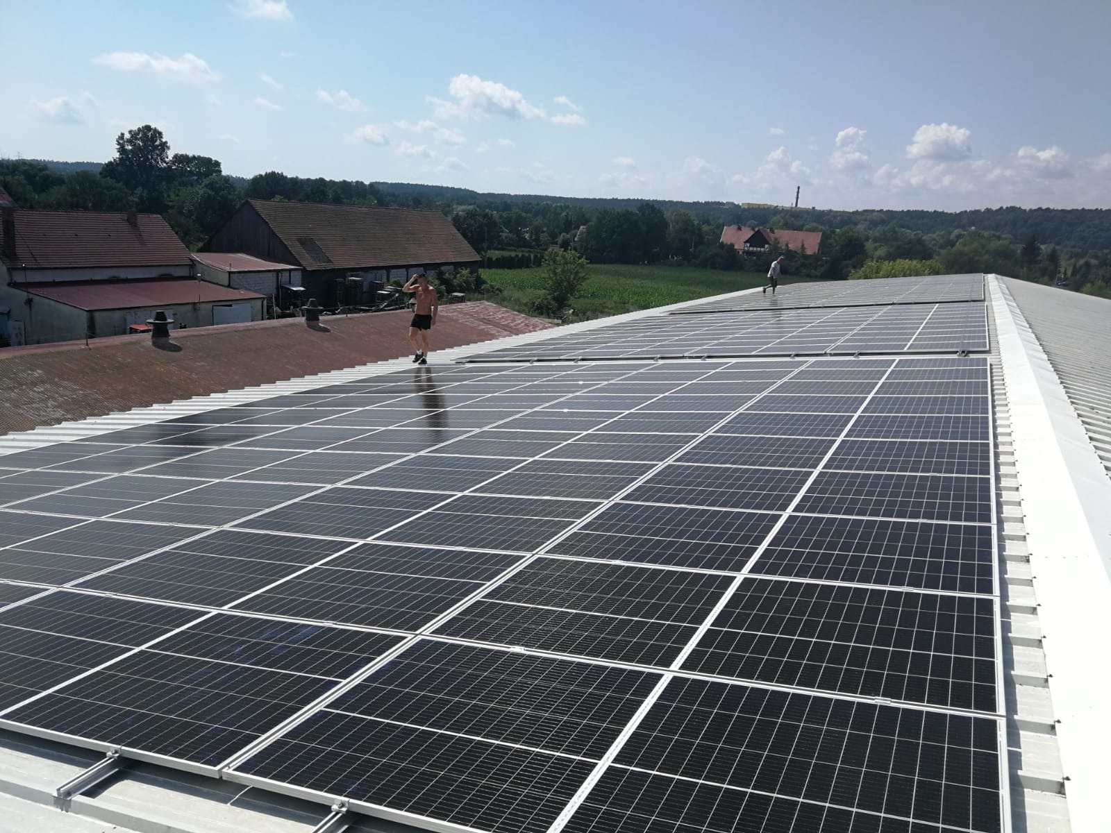 Montaż paneli fotowoltaicznych na dachu budynku gospodarczego z widocznymi pracownikami w oddali, otoczonym zielenią pól i lasów pod błękitnym niebem.