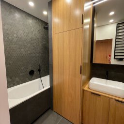 KOCHACZEWSKA DESIGN ANNA KOCHACZEWSKA-LIS - Nowoczesna łazienka z wanną obudowaną ciemnoszarym panelem, heksagonalnymi płytkami na ścianie, drewnianą zabudową meblową z szafką i umywalką nablatową oraz lustrem z oświetleniem LED.