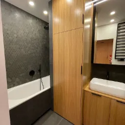 Nowoczesna łazienka z wanną obudowaną ciemnoszarym panelem, heksagonalnymi płytkami na ścianie, drewnianą zabudową meblową z szafką i umywalką nablatową oraz lustrem z oświetleniem LED.